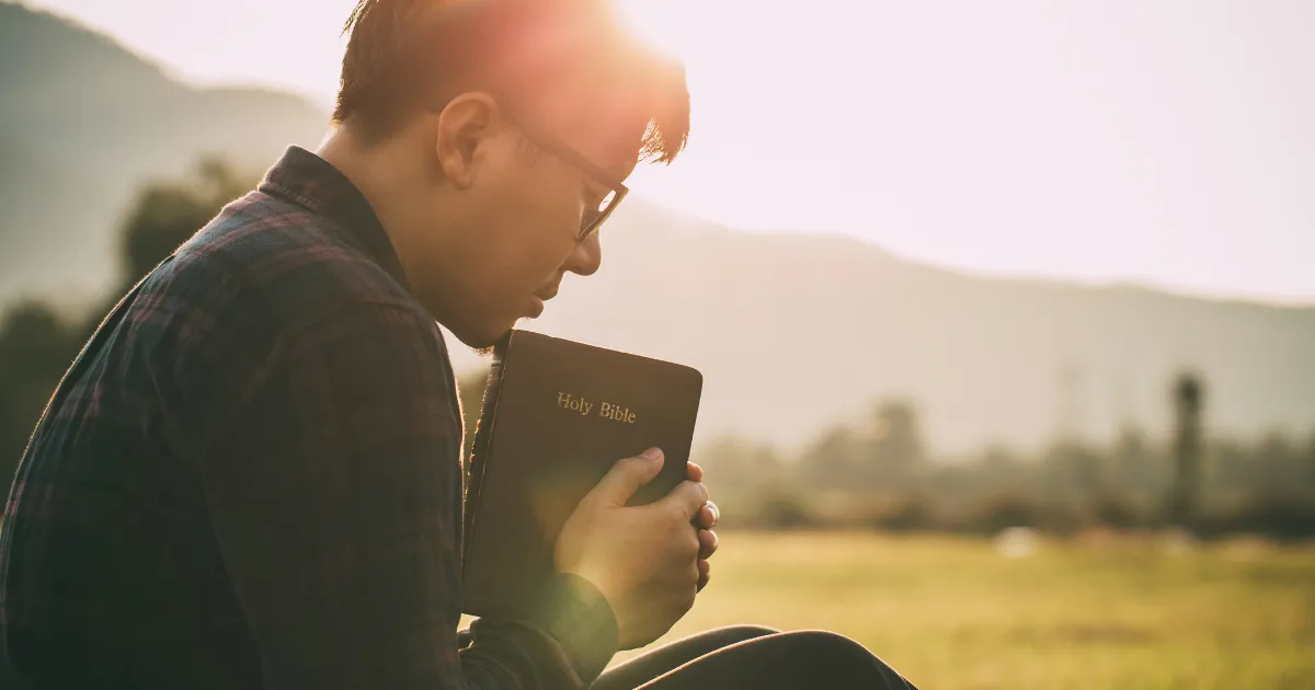 A man sitting peacefully in a field at sunrise, holding and reading the Holy Bible, representing spending quality time with God.
